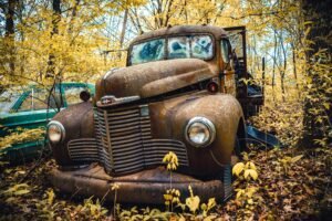 A vintage, rusty truck sits abandoned in a vibrant autumn forest, surrounded by yellow leaves.